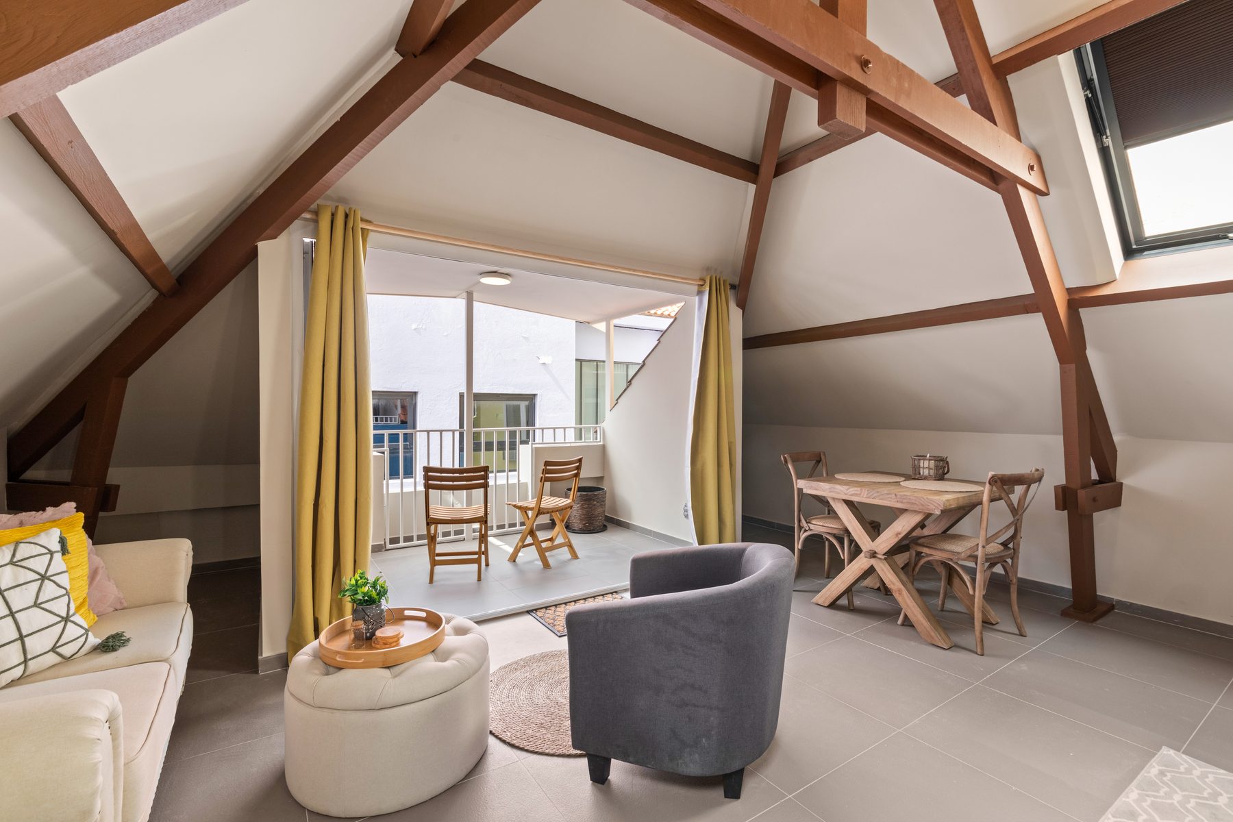 Sunlit loft living room beneath exposed timber roof trusses, opening onto a terrace